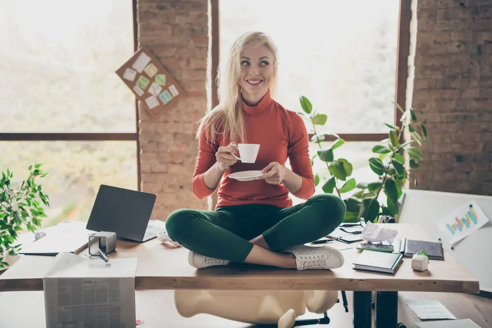 Full body photo of happy positive woman company owner sit on table, crossed legs rest relax hold coffee cup drink cappuccino wear red turtleneck green pants trousers in messy office loft